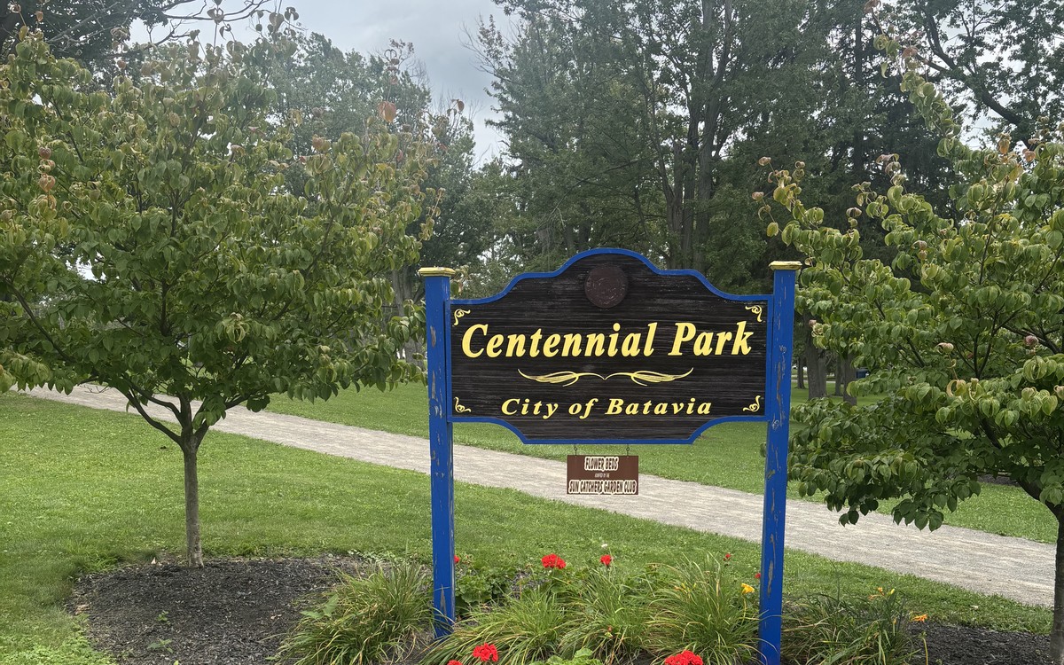 Centennial Park sign in Batavia with red flowers at its base and mature trees behind